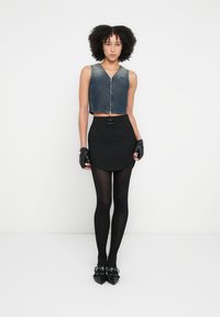 Džinsinė trumpa liemenė su priekiu užtrauktuku, derinama su juoda sijonu, kuriame yra išlenkta apatinė kraštinė, juodos pėdkelnės ir smailiomis juodomis batelėmis.