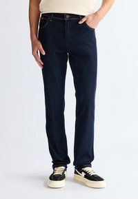 Mężczyzna noszący ciemnoniebieskie jeansy o prostych nogawkach, z jedną ręką w kieszeni oraz czarno-białe sznurowane trampki, stoi na białej podłodze.