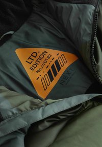 Zöld dzseki belseje fekete bolyhos béléssel, amelyen egy narancssárga háromszögletű címke található, rajta a "LTD EDITION No. 11/0182" és a "NEXT." felirattal.