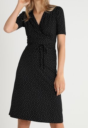 Woman wearing a black wrap dress with short sleeves and white polka dots, tied at the waist with a fabric belt.