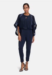 Navy jumpsuit with an overlay, featuring a keyhole neckline adorned with beads, flowing sleeves, and tapered legs. Black strappy heels complete the look.