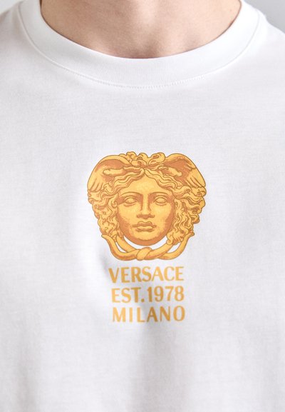 White shirt with a gold Versace Medusa logo above "VERSACE EST.1978 MILANO" text on chest.