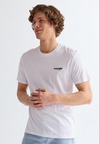 Ung mand med krøllet hår iført en hvid Wrangler t-shirt og lyseblå jeans, stående med hænderne foldet, kigger mod venstre.