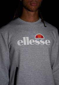 Sudadera gris con un logo blanco de "ellesse" y un acento en forma de medio círculo en rojo y naranja. Manga larga y cuello de canalé.