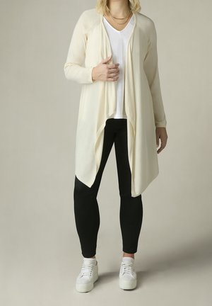 Personne portant un long cardigan crème, un T-shirt blanc, des leggings noirs, des baskets blanches et des colliers dorés superposés, debout devant un fond uni.