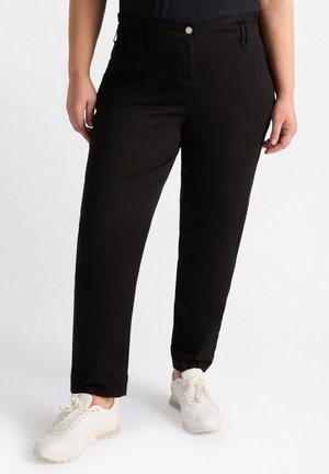 Vrouw die zwarte rechte broek met voorste knoop en witte sneakers draagt, staand tegen een effen witte achtergrond.