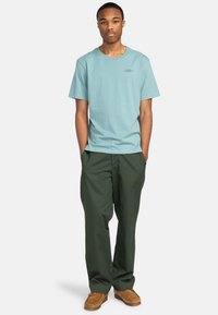 Hellblaues Baumwoll-T-Shirt mit Rundhalsausschnitt, kombiniert mit dunkelgrünen, locker sitzenden Hosen und braunen Slippers. Schlichter, funktioneller Look.