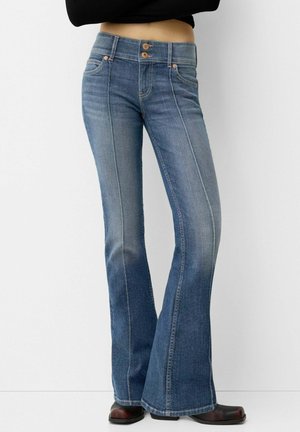Vrouw draagt een high waisted, blauwe uitlopende jeans met dubbele houten knopen en donkerbruine schoenen met vierkante neus, staand tegen een witte muur.