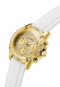 Montre dorée avec un cadran doré texturé, accents en cristal et un bracelet en silicone blanc. Elle dispose de trois sous-cadrans et d'une lunette proéminente.