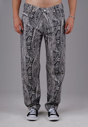 Mand iført sort- og hvidmønstrede bukser med sneakers, stående mod en ensfarvet grå baggrund.