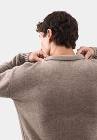 Jasnobrązowy sweter dzianinowy z okrągłym dekoltem i ściągaczami, o wyraźnej strukturze i miękkim materiale. Osoba poprawia dekolt.