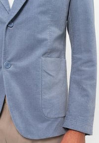 Blue corduroy blazer jacket with patch pocket worn over beige pants, showing buttons and sleeve on a person's arm.