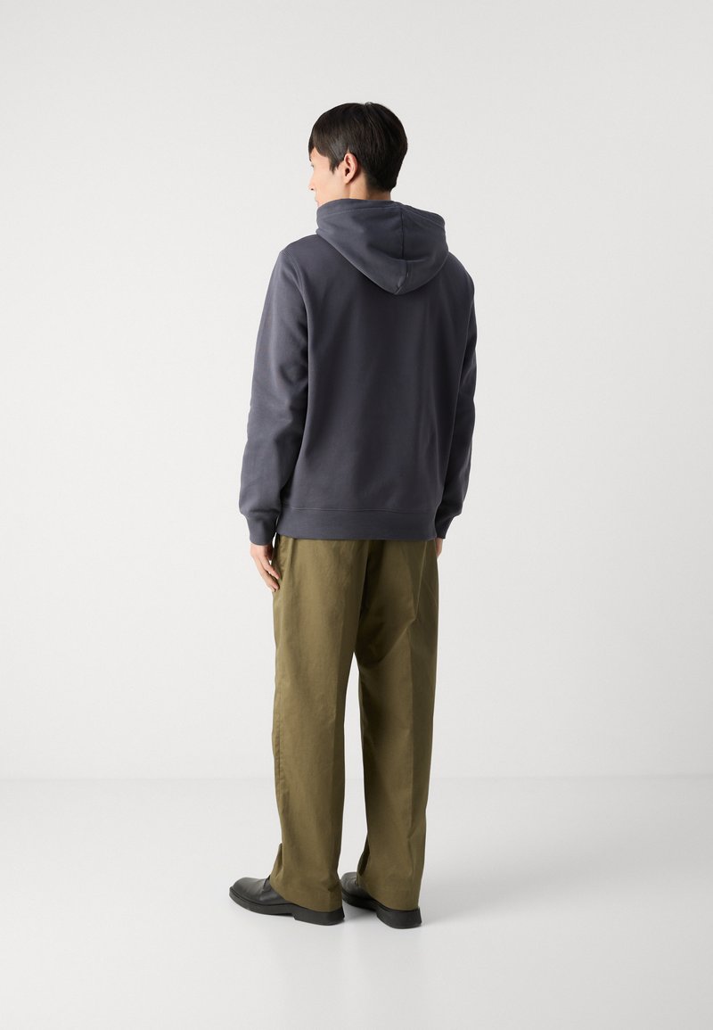 Sudadera con capucha en gris oscuro, con una tela texturizada. Combinada con pantalones sueltos de color verde oliva y zapatos negros, vista desde atrás.