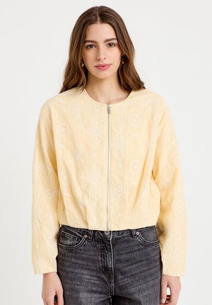 Young woman with long brown hair wears a light yellow floral zip-up jacket and dark gray jeans, standing against a white background.