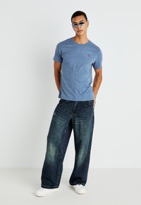 Μπλε βαμβακερό μπλουζάκι με λεπτή υφή και μικρό κόκκινο λογότυπο, συνδυασμένο με φαρδιά σκούρα τζιν παντελόνια και λευκά αθλητικά παπούτσια, σε φωτεινό φόντο.