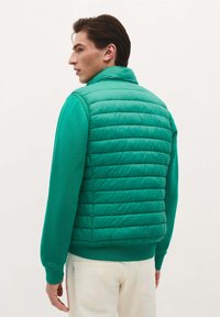 Chaleco acolchado verde con un alto cuello, que presenta segmentos acolchados. Llevado sobre una camisa de manga larga verde a juego, que resalta la textura suave.