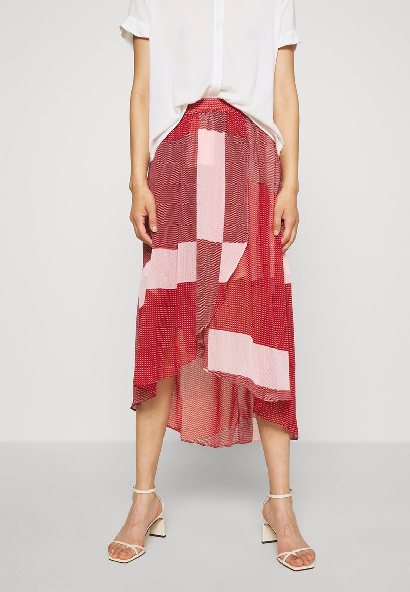 Jupe midi à motifs rouges et roses, avec un ourlet asymétrique, une texture transparente et une coupe décontractée. Associée à une chemise blanche et des sandales.