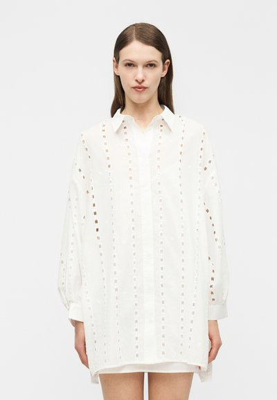 Femme portant une robe chemise blanche ample à manches longues avec des rayures découpées verticales, debout devant un fond uni.