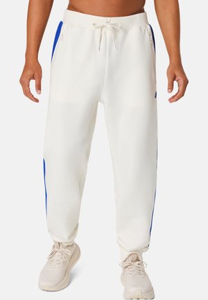 Pantaloni della tuta bianchi con pannelli laterali blu, tasche con zip, vita con coulisse, indossati con scarpe da ginnastica beige e torso inferiore e mani visibili.