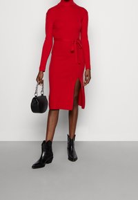 Robe en tricot rouge à manches longues, silhouette ajustée, fente sur le côté et taille à nouer. Associée à des bottes noires à la cheville et un petit sac à main noir.