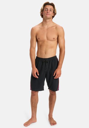 Shirtless young man with medium-length blond hair wearing black swim trunks with red side stripes standing barefoot against white background.
