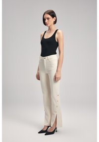 Pantaloni crema a vita alta con spacchi laterali e dettagli in bottone; abbinati a un body nero a coste e a tacchi neri a punta. 