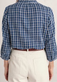 Chemise à carreaux bleue avec motif à carreaux blancs, encolure froncée et manches trois-quarts, associée à un pantalon clair avec poches arrière.