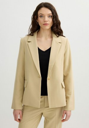 Mujer con cabello largo y ondulado, vistiendo una blazer beige sobre una blusa negra y pantalones beige a juego, de pie contra un fondo claro y liso.