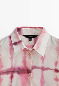 Chemise à col avec boutons et motif tie-dye rose et violet sur tissu blanc, montrant le haut devant et le détail du col.