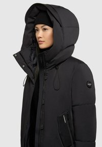Sort isoleret parka med en stor hætte, knap- og lynlåslukninger, elastiske snore samt en lynlåslomme på siden. Blød tekstur.