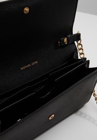 Černá syntetická kabelka se zlatým kováním, obsahující řetízkový popruh a více vnitřních přihrádek. Vryté detaily "MICHAEL KORS".
