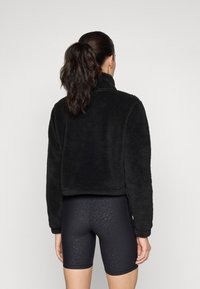 Veste en polaire noire cropped avec un col haut, présentant une texture douce. Associée à des shorts de vélo noirs affichant un motif léopard subtil.