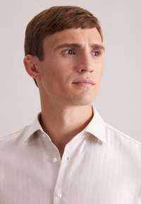 Camicia button-up color beige chiaro con texture a spina di pesce, colletto e chiusura con un solo bottone sul davanti, indossata da un uomo con espressione neutra.