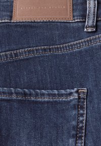 Jeans de mezclilla azul oscuro con una textura suave, costuras en contraste y una etiqueta de cuero marrón etiquetada "STREET ONE STUDIO" en la cintura.