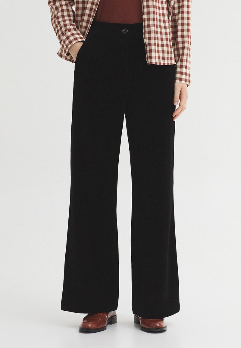Personne portant un pantalon noir à jambes larges, des chaussures marron, une chemise rouge et une veste rouge et blanche à carreaux, debout devant un fond clair.