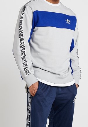 Mann trägt ein hellgraues Sweatshirt mit einem blauen Brustpaneel und gemusterten schwarz-weißen Streifen an den Ärmeln sowie marineblaue Jogginghose mit den Händen in den Taschen.