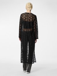 Cappotto lungo in pizzo nero con maniche lunghe, schiena trasparente e motivo floreale, che si estende sotto il ginocchio. Indossato sopra pantaloni grigi e scarpe.