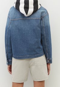 Giacca di denim di colore blu medio con cappuccio a righe bianche e nere. Presenta polsini con bottone e una vestibilità rilassata. Abbinata a pantaloni corti beige.
