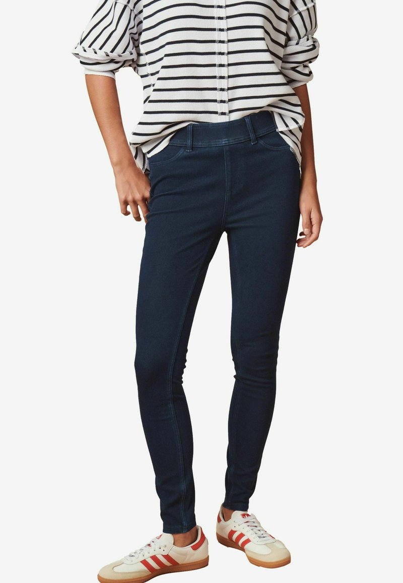 Mörkblå skinny jeans med en slät textur, hög midja och sidofickor, i kombination med en vit och svart randig topp och vita sneakers.