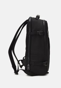 Sac à dos rectangulaire noir avec des bretelles rembourrées, plusieurs zips, des boucles sur les côtés et une poignée sur le dessus, sur fond blanc.