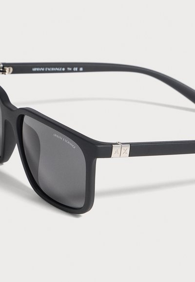 Lunettes de soleil noires Armani Exchange, à monture rectangulaire, verres gris foncé et détails AX argentés sur les branches, sur fond clair.