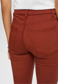 Personne portant un jean ajusté couleur rouille avec des poches arrière et un haut blanc, montrée de la taille au milieu de la cuisse.