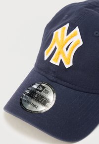 Șapcă albastru închis cu curea reglabilă, având un logo NY brodat galben pe partea din față și o etichetă circulară neagră New Era pe vizieră.