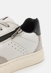 Világosszürke sneaker velúr részletekkel, perforált elemekkel és fekete sarokkal. Oldalsó cipzárral és fűzőzáródással rendelkezik. A márka logója látható.