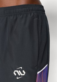 Μαύρες αθλητικές βερμούδες με διαπνέουσες πλαι�νές επενδύσεις, ελαστική μέση και μοβ διαβαθμισμένα στοιχεία με λεπτομέρεια λευκού λογότυπου.