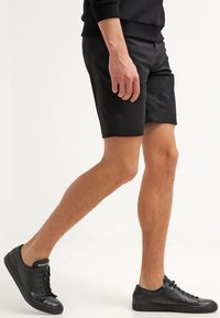 Schwarze Shorts aus glattem Stoff, mit taillierter Passform und Seitentaschen. Kombiniert mit schwarzen Sneakers für einen harmonischen Look.