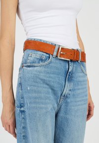 Brun, texturerad bälte med en silverfärgad metallspänne, som bärs med ljusblå, högmidjade jeans och en vit åtsittande topp.