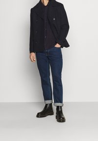 Sort uld dobbeltbrystet jakke over en mørk skjorte, parret med mørkeblå jeans med op-rullede ben og sorte ankelstøvler med en kraftig sål.