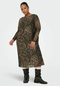 Donna che indossa un vestito midi a maniche lunghe con stampa leopardo, stivali marrone scuro fino al polpaccio e calzini bianchi, in piedi davanti a uno sfondo neutro.