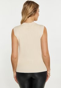 Top corto a maniche corte color beige chiaro, con texture a costine, design aderente e scollo rotondo. Indossato con pantaloni neri lucidi a vita alta.